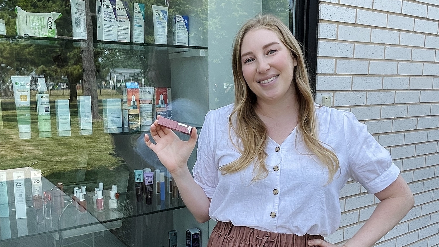 Mira Abed stands to the left of the image holding a Maybelline Vinyl Ink lip product. Behind Abed is a product window display at the L'Oreal New Jersey location.