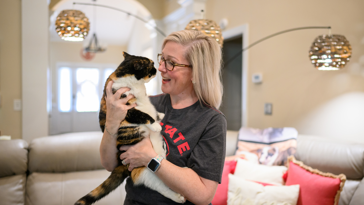 A woman with blonde, shoulder-length hair wearing a gray NC State t-shirt smiles at the calico cat she is holding in her living room.