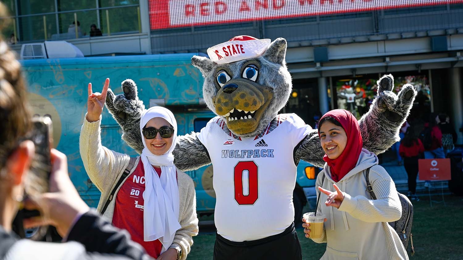 Two students standing on either side of the NC State mascot, making the Wolfpack sign.